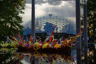 A vibrant, multicolored glass sculpture displayed on a reflective water surface in front of a glass-domed conservatory. The sky above is partly cloudy with rays of sunlight streaming through. People can be seen walking around in the background, and tall trees frame the scene.