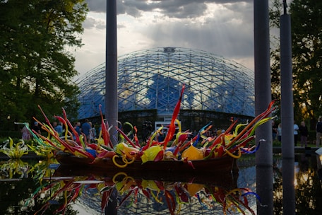 A vibrant, multicolored glass sculpture displayed on a reflective water surface in front of a glass-domed conservatory. The sky above is partly cloudy with rays of sunlight streaming through. People can be seen walking around in the background, and tall trees frame the scene.