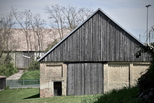 A rustic wooden barn stands prominently, with weathered wooden planks forming the front facade and a sloped roof. The barn is set against a backdrop of sparse trees and another building with a brick pattern visible behind it. In the foreground, green grass and some bushes add natural elements to the scene.
