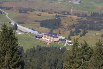 A large building with a dark roof surrounded by lush green fields and dense forests. A network of roads and paths can be seen connecting the building with the surrounding area, with a nearby parking lot and several parked cars. The landscape features rolling hills and scattered trees, creating a serene and scenic atmosphere.
