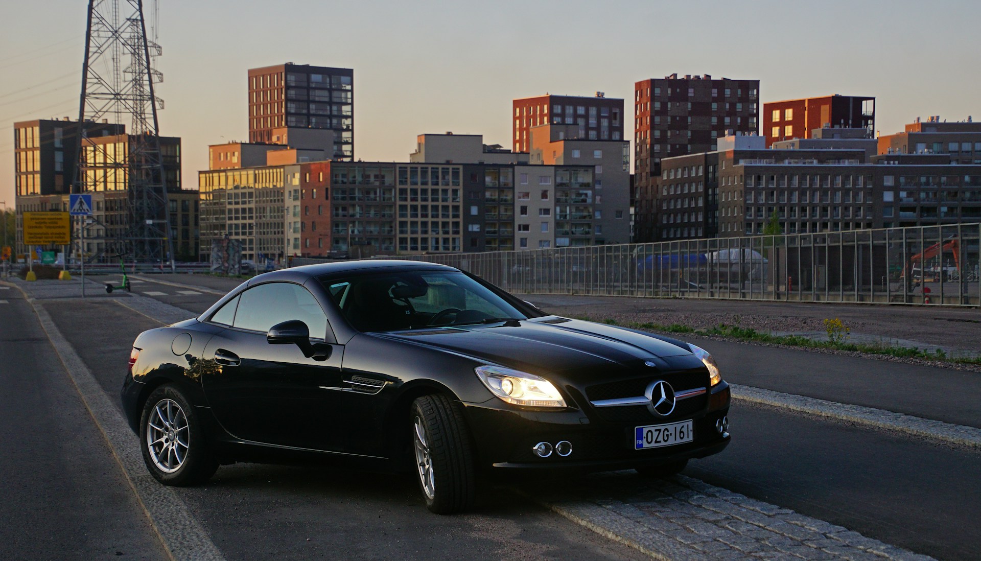A luxury black convertible with the top down, parked in front of a modern city skyline at sunset.
