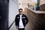 A person wearing sunglasses and a black jacket with 'NEW YORK' written on it stands in an urban alleyway. The background features modern architectural elements, including concrete and glass buildings.