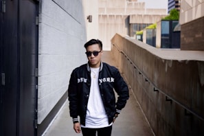 A person wearing sunglasses and a black jacket with 'NEW YORK' written on it stands in an urban alleyway. The background features modern architectural elements, including concrete and glass buildings.