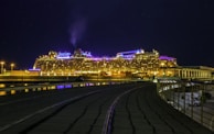 A large cruise ship is illuminated with bright lights against a dark night sky. The ship is docked at a port, with visible pathways and a railing in the foreground. The lights reflect off the water, creating a vibrant and lively atmosphere.