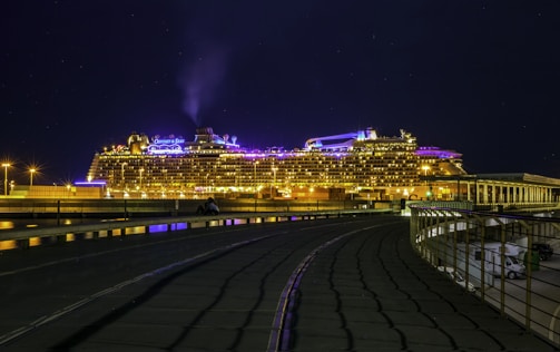 A large cruise ship is illuminated with bright lights against a dark night sky. The ship is docked at a port, with visible pathways and a railing in the foreground. The lights reflect off the water, creating a vibrant and lively atmosphere.