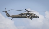 A military helicopter is flying in the sky with several soldiers visible inside. The aircraft has dark green camouflage, and the soldiers are equipped with gear, appearing ready for operation. The sky is mostly clear with some white clouds.