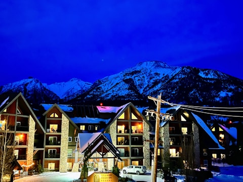 Chalet-style buildings with illuminated windows, set against a backdrop of snow-capped mountains under a deep blue sky. A parked vehicle and snow-covered ground are visible in the foreground with a lit sign reading 'GRANDE ROCKIES'.