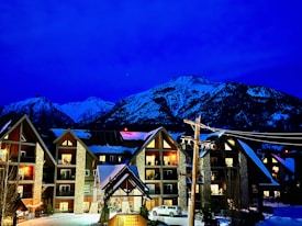 Chalet-style buildings with illuminated windows, set against a backdrop of snow-capped mountains under a deep blue sky. A parked vehicle and snow-covered ground are visible in the foreground with a lit sign reading 'GRANDE ROCKIES'.