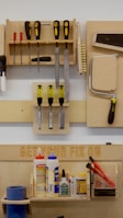 A neatly organized wall-mounted tool rack featuring various hand tools such as screwdrivers, files, brushes, and a saw. Below, there is a shelf with numerous glue bottles, adhesives, and a pair of red-handled pliers. The phrase 'GET YOUR FIX ON' is engraved on a wooden board above the shelf.