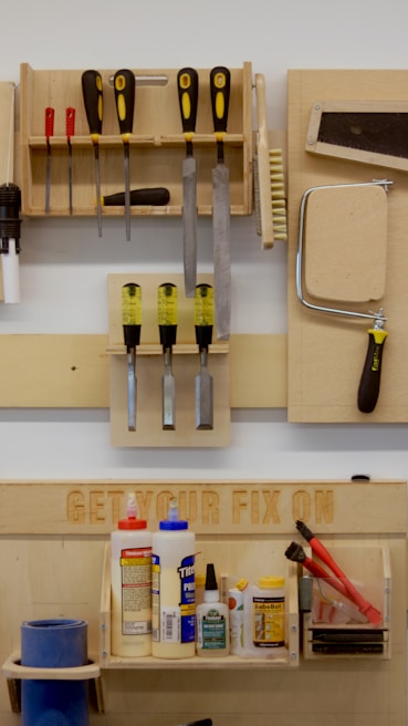 A neatly organized wall-mounted tool rack featuring various hand tools such as screwdrivers, files, brushes, and a saw. Below, there is a shelf with numerous glue bottles, adhesives, and a pair of red-handled pliers. The phrase 'GET YOUR FIX ON' is engraved on a wooden board above the shelf.