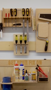 A neatly organized wall-mounted tool rack featuring various hand tools such as screwdrivers, files, brushes, and a saw. Below, there is a shelf with numerous glue bottles, adhesives, and a pair of red-handled pliers. The phrase 'GET YOUR FIX ON' is engraved on a wooden board above the shelf.