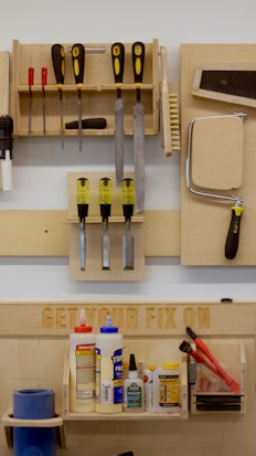 A neatly organized wall-mounted tool rack featuring various hand tools such as screwdrivers, files, brushes, and a saw. Below, there is a shelf with numerous glue bottles, adhesives, and a pair of red-handled pliers. The phrase 'GET YOUR FIX ON' is engraved on a wooden board above the shelf.