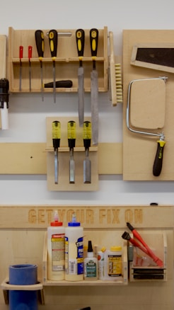 A neatly organized wall-mounted tool rack featuring various hand tools such as screwdrivers, files, brushes, and a saw. Below, there is a shelf with numerous glue bottles, adhesives, and a pair of red-handled pliers. The phrase 'GET YOUR FIX ON' is engraved on a wooden board above the shelf.
