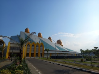 The architecture features a modern structure with a series of golden domes and a distinct geometric design. Palm trees are situated along a paved road leading up to the building, creating a welcoming environment. The facade includes large, arching windows accented with yellow trim.