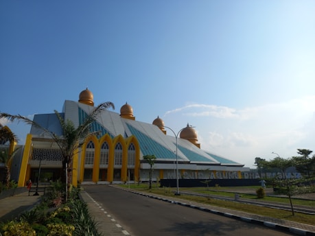 The architecture features a modern structure with a series of golden domes and a distinct geometric design. Palm trees are situated along a paved road leading up to the building, creating a welcoming environment. The facade includes large, arching windows accented with yellow trim.