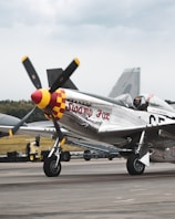 A vintage aircraft is parked on a runway, with the name 'Swamp Fox' painted on its metallic body. The plane features a propeller with a red and yellow checkered nose. In the cockpit, a pilot is sitting with one hand raised, wearing a helmet and sunglasses. The background shows another aircraft's tail and a grassy field under a cloudy sky.