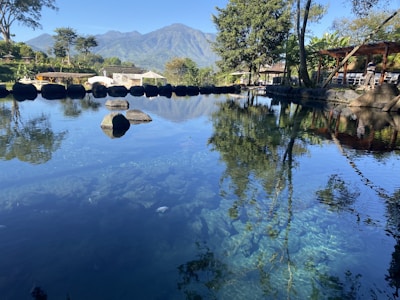 A calm, clear pond reflects the surrounding trees and distant mountains under a blue sky. There are several rocks scattered along the pond's edge, and lush greenery surrounds the area. In the background, a structure with a thatched roof is partially visible.