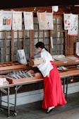 A person dressed in traditional attire is handling items in trays at a stall or shop. Above the stall are various posters with text and illustrations, some listing prices in yen. The setting appears to be indoors with wood-paneled doors in the background.