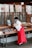 A person dressed in traditional attire is handling items in trays at a stall or shop. Above the stall are various posters with text and illustrations, some listing prices in yen. The setting appears to be indoors with wood-paneled doors in the background.