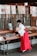 A person dressed in traditional attire is handling items in trays at a stall or shop. Above the stall are various posters with text and illustrations, some listing prices in yen. The setting appears to be indoors with wood-paneled doors in the background.
