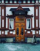 An ornate wooden door with intricate carvings is centered between two windows with white lace curtains. The building facade features a mix of white and reddish-brown wood paneling with decorative elements. A small black canopy extends over the door, and there is a menu stand on the right side.