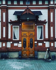 An ornate wooden door with intricate carvings is centered between two windows with white lace curtains. The building facade features a mix of white and reddish-brown wood paneling with decorative elements. A small black canopy extends over the door, and there is a menu stand on the right side.