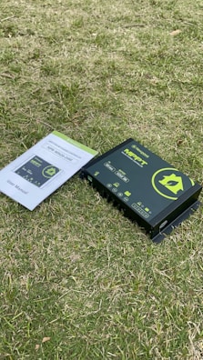 A solar charge controller with the brand Newpowa and model MPPT is placed on a grassy surface alongside its user manual. The charge controller features a green house icon, various symbols, and text describing its functions. The user manual is open to the front page, visible with green and white text.