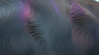 The image showcases a wavy, textured surface with flowing, organic lines and grooves. The surface has a three-dimensional appearance with gentle curves that create a sense of movement. A subtle blend of colors, including shades of purple and grey, adds depth to the abstract design.