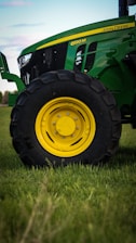 A close-up view of a green tractor parked on a grassy field, showing a large black wheel with a bright yellow rim. The tractor's body features a sleek design with visible branding and engine details.