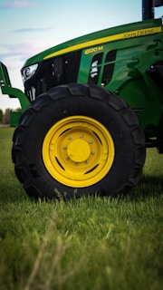 A close-up view of a green tractor parked on a grassy field, showing a large black wheel with a bright yellow rim. The tractor's body features a sleek design with visible branding and engine details.