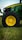 A close-up view of a green tractor parked on a grassy field, showing a large black wheel with a bright yellow rim. The tractor's body features a sleek design with visible branding and engine details.