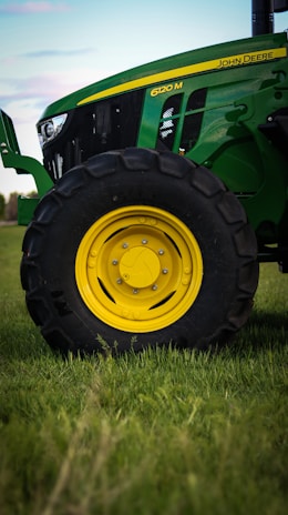 A close-up view of a green tractor parked on a grassy field, showing a large black wheel with a bright yellow rim. The tractor's body features a sleek design with visible branding and engine details.