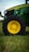 A close-up view of a green tractor parked on a grassy field, showing a large black wheel with a bright yellow rim. The tractor's body features a sleek design with visible branding and engine details.