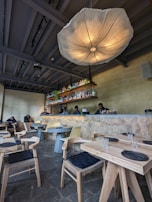 The interior of a restaurant with modern, minimalistic decor. A large, artistic light fixture hangs prominently from the ceiling, illuminating the space. The bar area features a stone counter with bottles neatly arranged on shelves behind it. Several tables and chairs are neatly arranged in the foreground, each set with black cushioned seats and simple table settings. Three staff members stand behind the counter, suggesting a welcoming and friendly atmosphere.