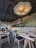 The interior of a restaurant with modern, minimalistic decor. A large, artistic light fixture hangs prominently from the ceiling, illuminating the space. The bar area features a stone counter with bottles neatly arranged on shelves behind it. Several tables and chairs are neatly arranged in the foreground, each set with black cushioned seats and simple table settings. Three staff members stand behind the counter, suggesting a welcoming and friendly atmosphere.