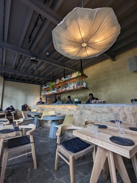 The interior of a restaurant with modern, minimalistic decor. A large, artistic light fixture hangs prominently from the ceiling, illuminating the space. The bar area features a stone counter with bottles neatly arranged on shelves behind it. Several tables and chairs are neatly arranged in the foreground, each set with black cushioned seats and simple table settings. Three staff members stand behind the counter, suggesting a welcoming and friendly atmosphere.