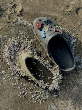 A single black Croc shoe adorned with colorful pins is placed on a sandy surface. Next to it, an imprint in the sand reveals another Croc shoe, heavily encrusted with barnacles. The texture of the sand and the barnacle-covered shoe frame create a contrast between the shoe's artificial material and the natural elements.