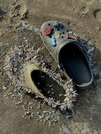 A single black Croc shoe adorned with colorful pins is placed on a sandy surface. Next to it, an imprint in the sand reveals another Croc shoe, heavily encrusted with barnacles. The texture of the sand and the barnacle-covered shoe frame create a contrast between the shoe's artificial material and the natural elements.