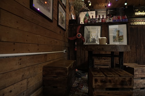 A cozy interior featuring wooden walls and furniture, adorned with colorful framed pictures. A round table with a wicker basket on top and square stools are placed in the corner. A vintage-style fan is situated near decorative bottles and plants, illuminated by ambient string lights.