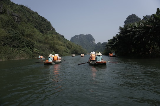 Rowboats filled with people wearing conical hats and orange life vests navigate a calm river. The river is flanked by lush green vegetation and towering limestone karsts under a clear blue sky.