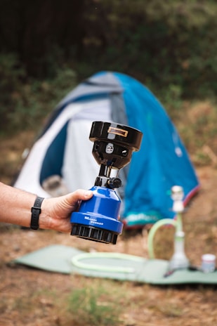 A hand holds a small portable gas burner with a blue base, labeled 'Wando Burner.' In the background, a blue and white tent is set up on a grassy patch with some camping equipment, including what appears to be a hookah, scattered around.
