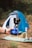 A hand holds a small portable gas burner with a blue base, labeled 'Wando Burner.' In the background, a blue and white tent is set up on a grassy patch with some camping equipment, including what appears to be a hookah, scattered around.