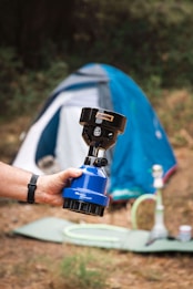 A hand holds a small portable gas burner with a blue base, labeled 'Wando Burner.' In the background, a blue and white tent is set up on a grassy patch with some camping equipment, including what appears to be a hookah, scattered around.
