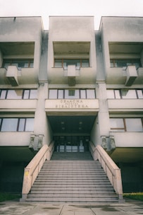 A large, Brutalist-style building with a prominent staircase leading to the entrance. The facade features geometric shapes and window arrangements. The sign above the entrance is in Cyrillic script.