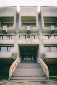 A large, Brutalist-style building with a prominent staircase leading to the entrance. The facade features geometric shapes and window arrangements. The sign above the entrance is in Cyrillic script.