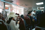 Inside a ferry or public transport vehicle with passengers seated and some standing. The interior has bright lighting and white fixtures, with a sense of packed seating arrangements. Many passengers are casually dressed and some are engaged with their phones.