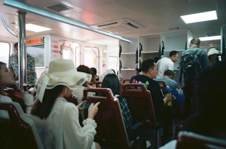 Inside a ferry or public transport vehicle with passengers seated and some standing. The interior has bright lighting and white fixtures, with a sense of packed seating arrangements. Many passengers are casually dressed and some are engaged with their phones.