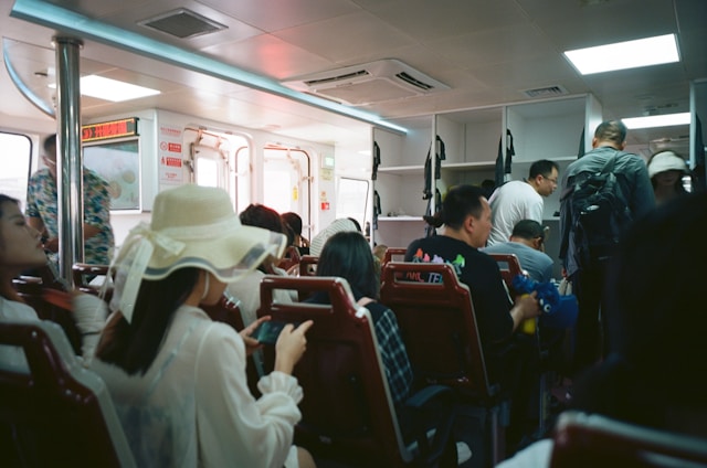 Inside a ferry or public transport vehicle with passengers seated and some standing. The interior has bright lighting and white fixtures, with a sense of packed seating arrangements. Many passengers are casually dressed and some are engaged with their phones.