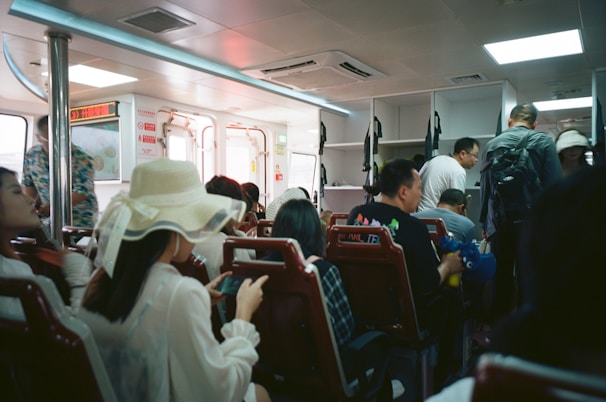 Inside a ferry or public transport vehicle with passengers seated and some standing. The interior has bright lighting and white fixtures, with a sense of packed seating arrangements. Many passengers are casually dressed and some are engaged with their phones.