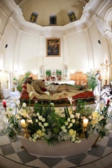 A grand religious interior features ornate architecture with high ceilings and large columns. At the center, a representation of Jesus lies in repose, surrounded by lush floral arrangements and glowing candles. Artwork depicting a saint is prominently displayed above.